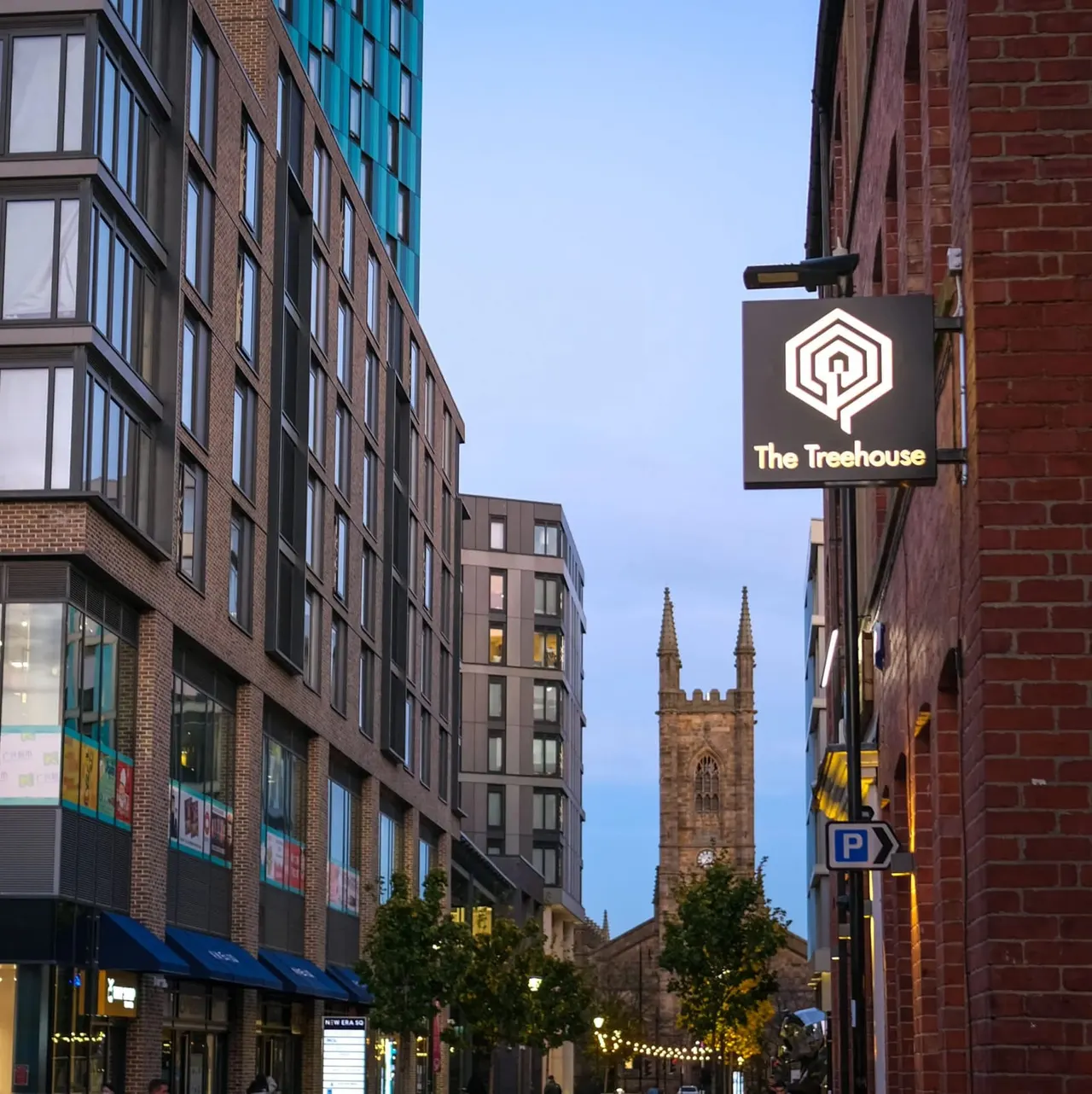 Urban street scene at dusk with The Treehouse venue sign, historic stone church spire visible between brick buildings.