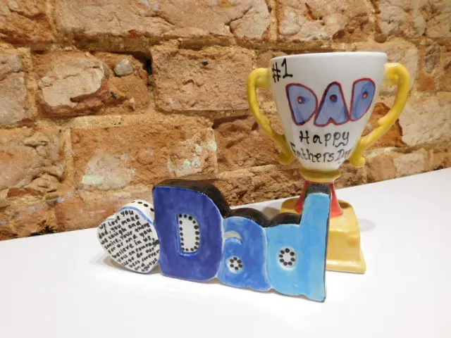 Handmade ceramic pieces spelling "DAD" in blue tones alongside a trophy-style mug against a rustic brick wall.