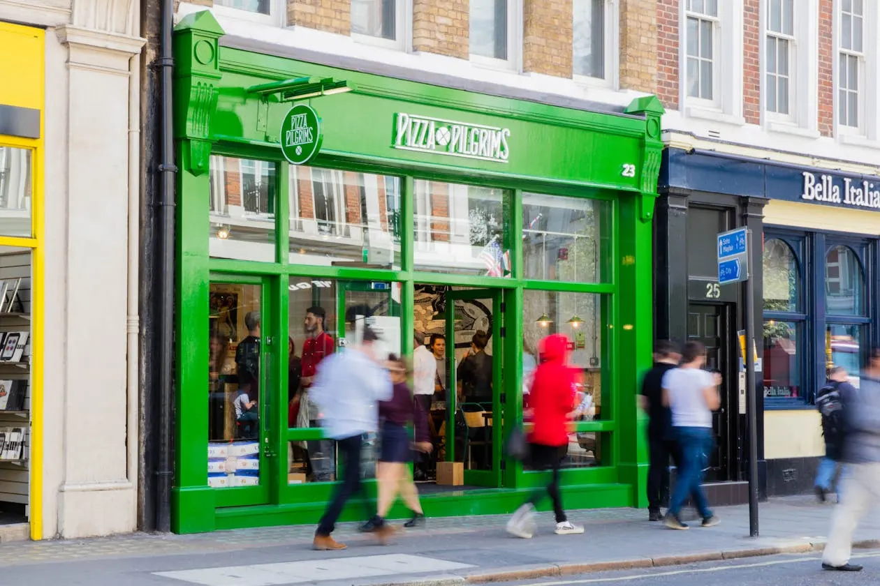 Pedestrians walking past a bright green-fronted Pizza Pilgrims restaurant on a busy street.