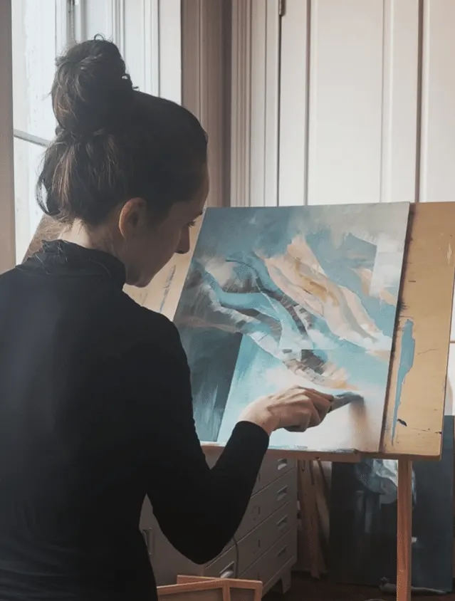 Woman painting abstract artwork on canvas at an easel near bright windows.