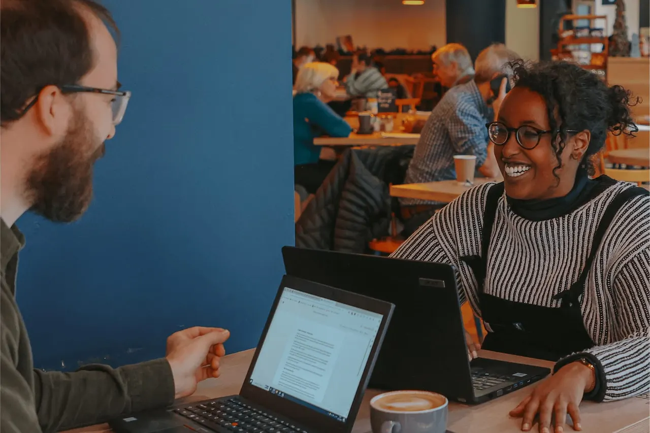 Two people sit at a wooden table with laptops, smiling whilst having a conversation in a busy café.