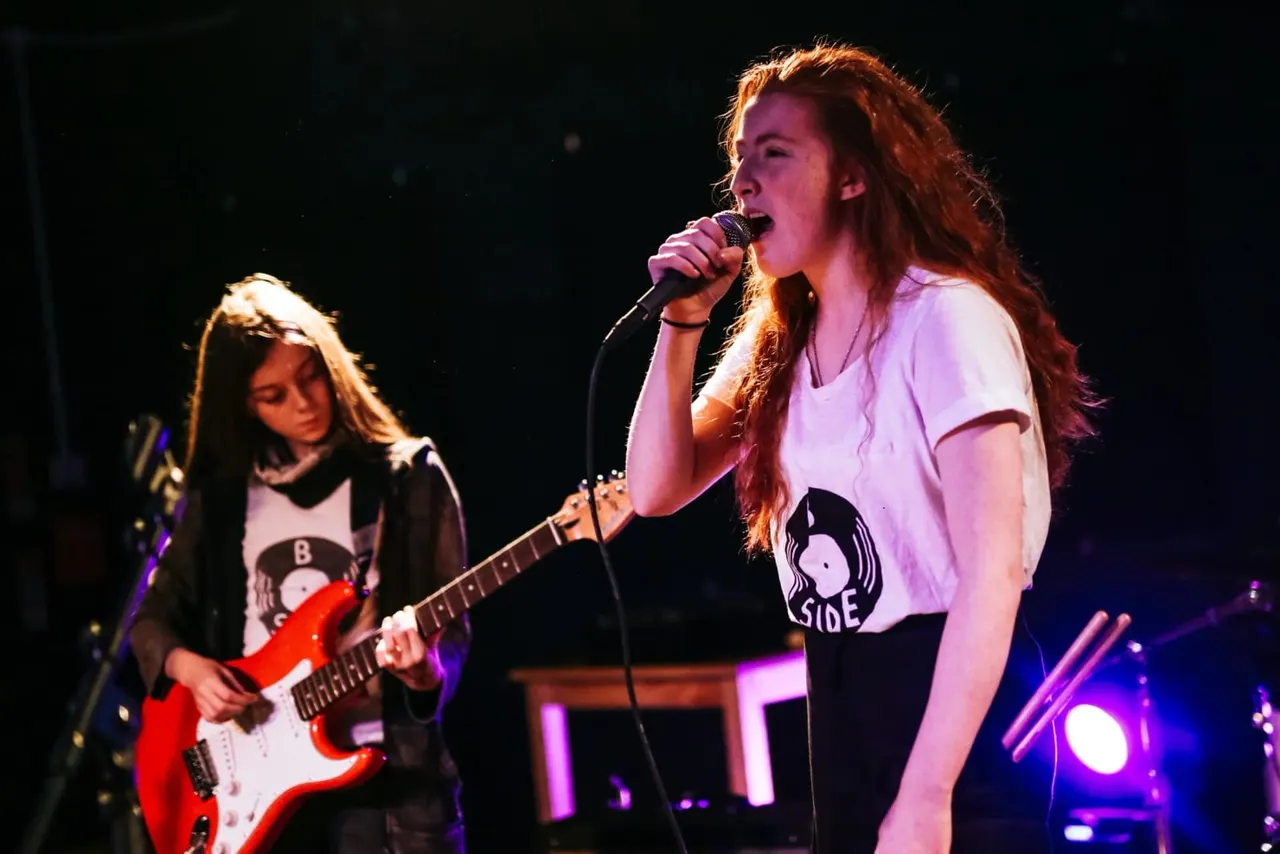 Two female musicians performing on stage; one singing into a microphone, the other playing a red electric guitar.