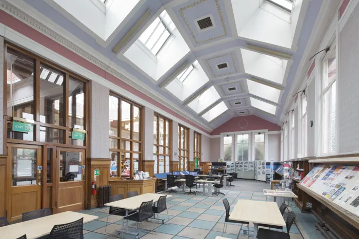 Interior of a bright library reading room with skylights, wooden doors, tables, chairs and bookshelves.