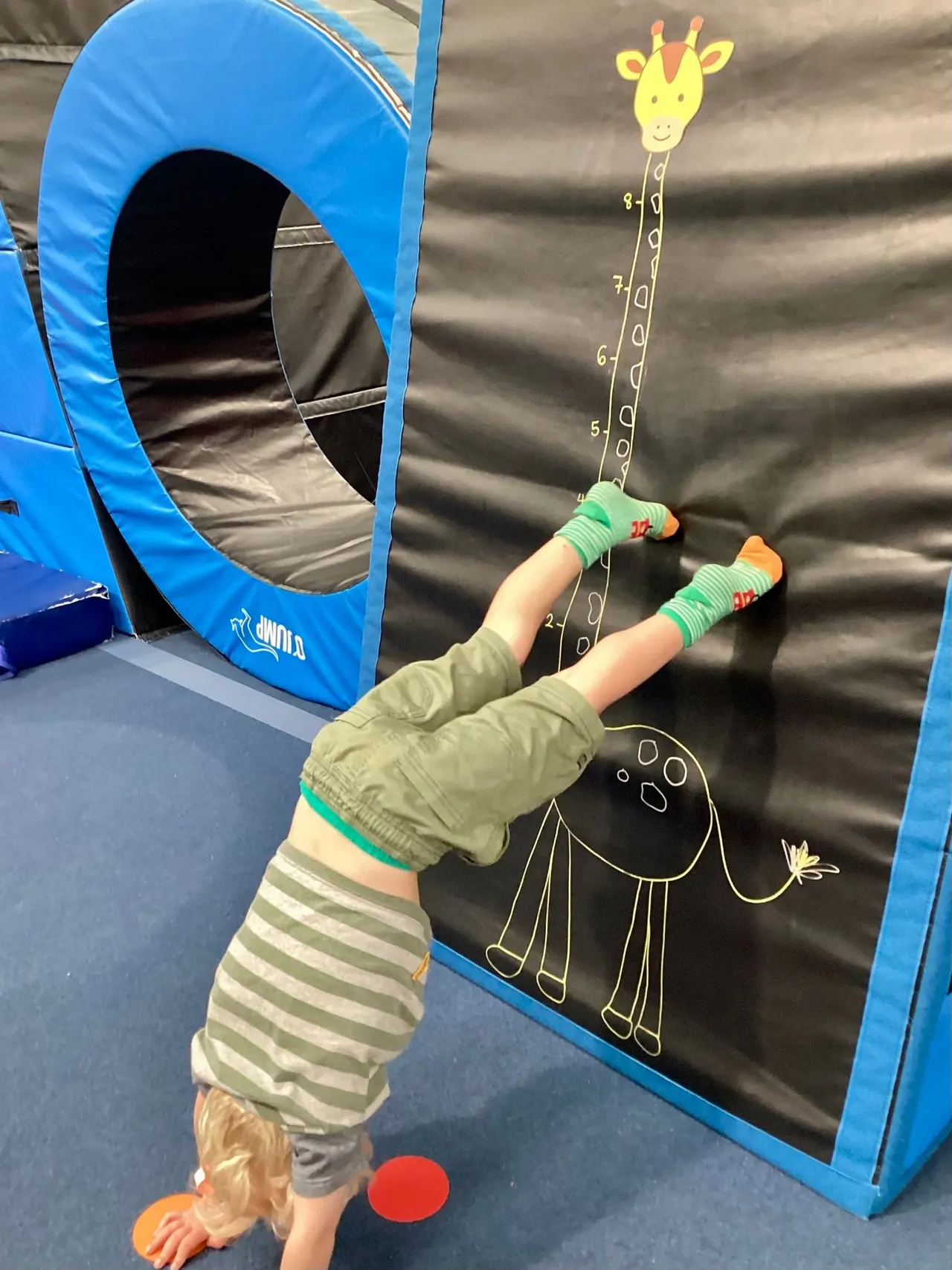 Child doing a handstand on a trampoline next to a blue tunnel, reaching towards a height chart with a giraffe illustration.