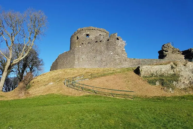 Ruins of a stone castle with a round tower on a grassy mound under blue sky, with bare trees nearby.