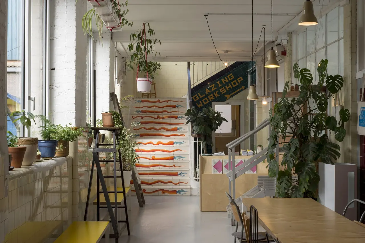 Bright studio space with potted plants hanging and on windowsills, a decorated staircase with orange patterns, wooden tables, and a "Glazing Workshop" sign.