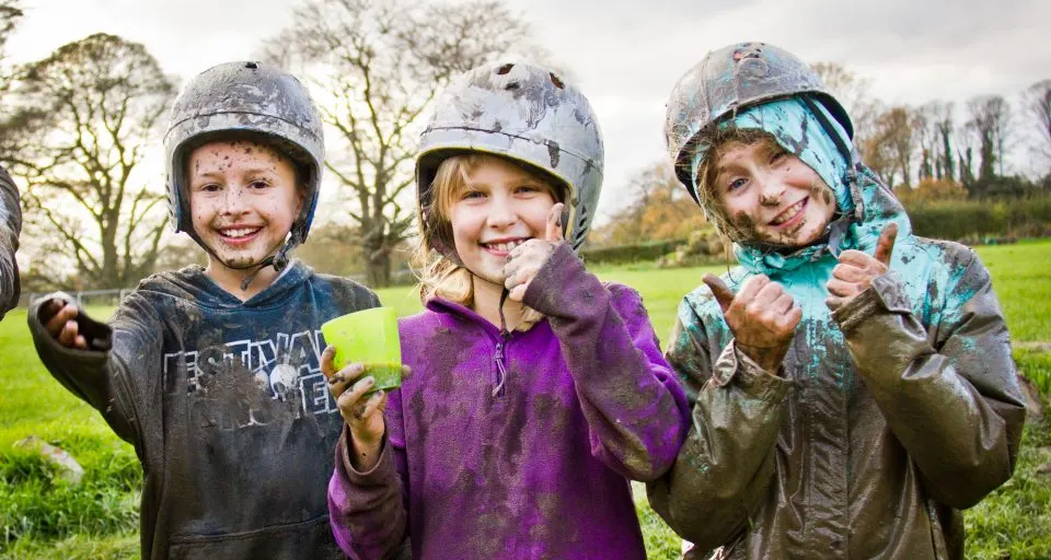 Three young children wearing protective helmets stand in a grassy field, smiling and giving thumbs up.