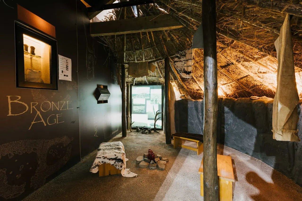 Interior of a Bronze Age museum exhibit with thatched roof, wooden posts, animal hide, firepit and informational displays.