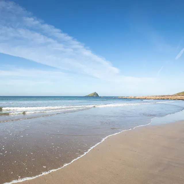 Sandy beach with gentle waves, rocky headland in distance under clear blue sky.