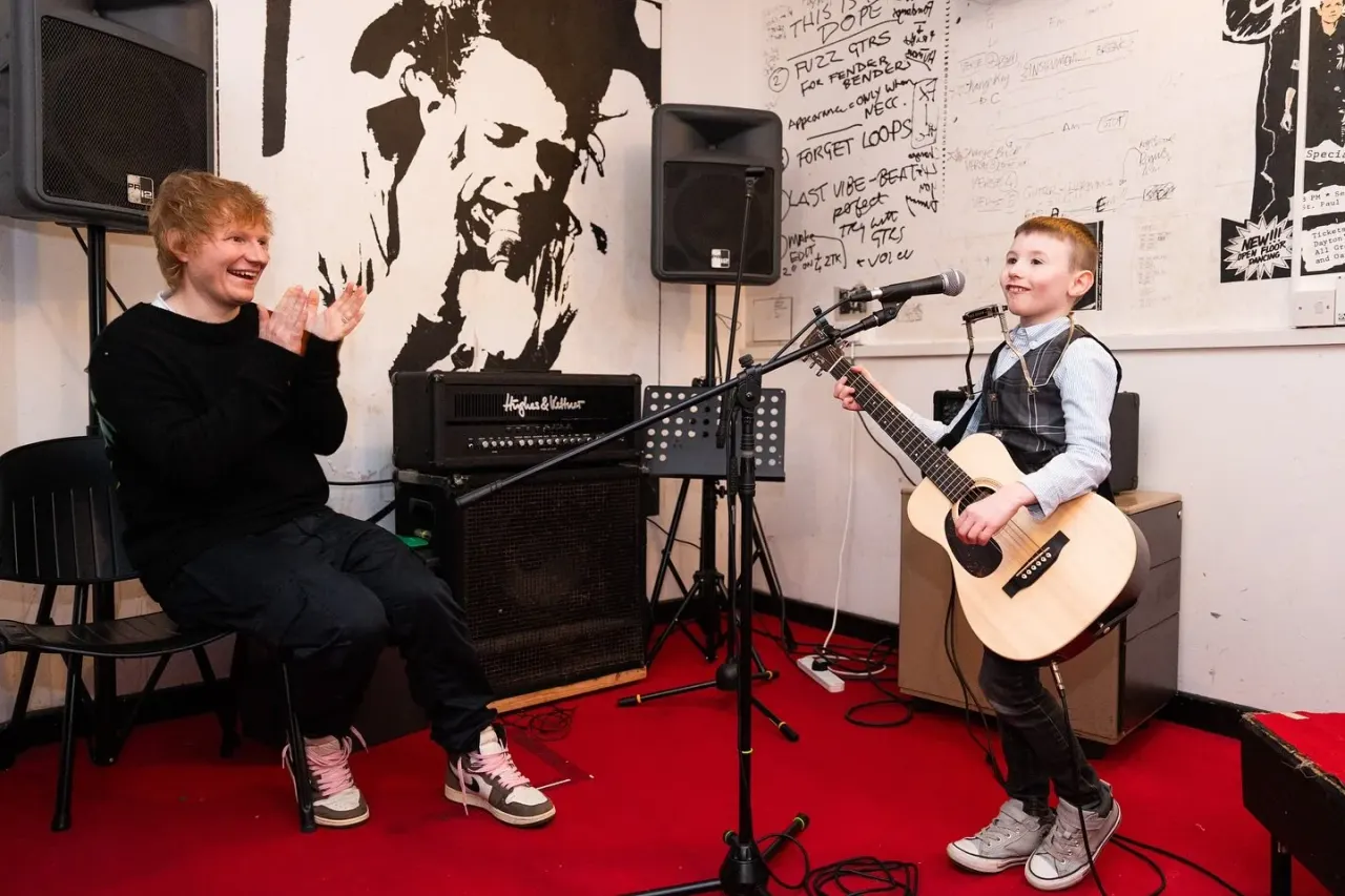 Two young children performing in a music studio, one playing acoustic guitar whilst the other sits nearby clapping.