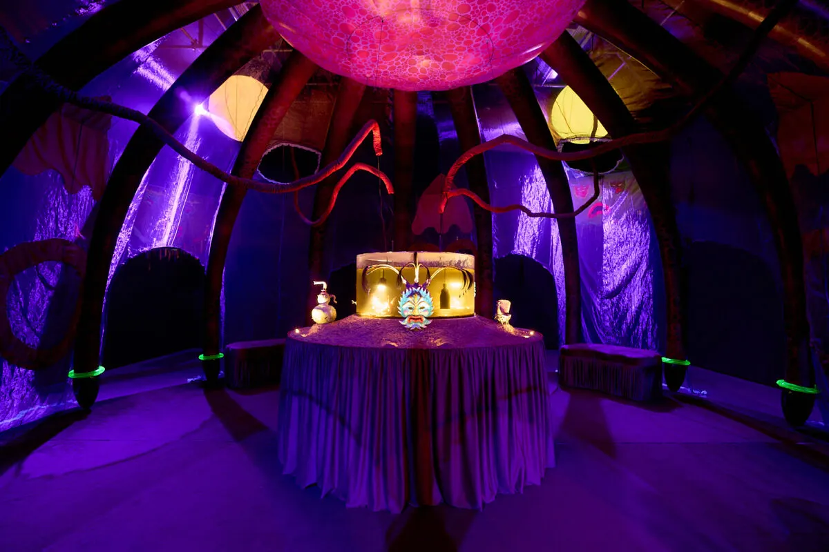 Colourful illuminated interior space with purple, pink and yellow lighting, a central draped table display, and neon signage in a curved architectural setting.
