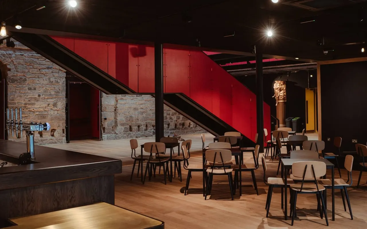 Modern theatre bar with wooden floors, tables, chairs, exposed stone walls and striking red staircase under dramatic lighting.