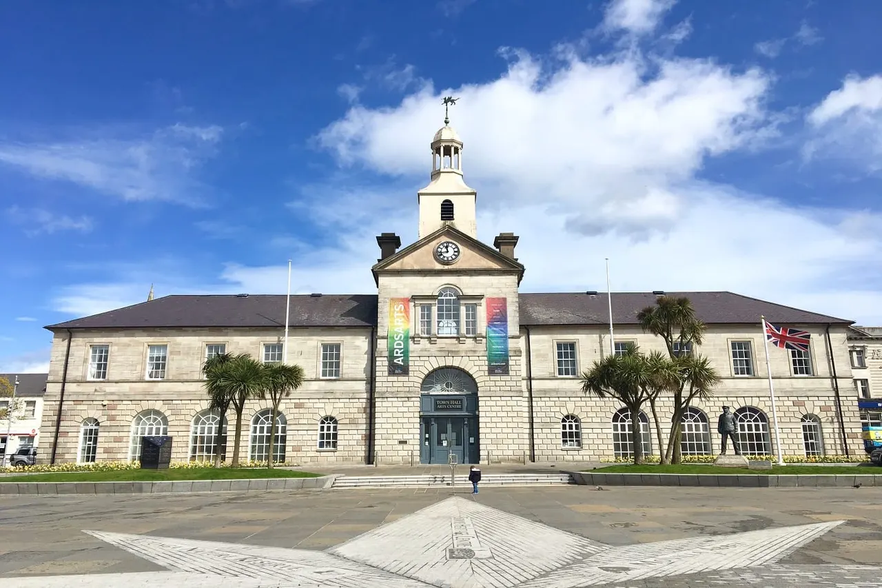 Historic stone building with clock tower and cupola, flanked by palm trees, viewed across a paved forecourt on a sunny day.