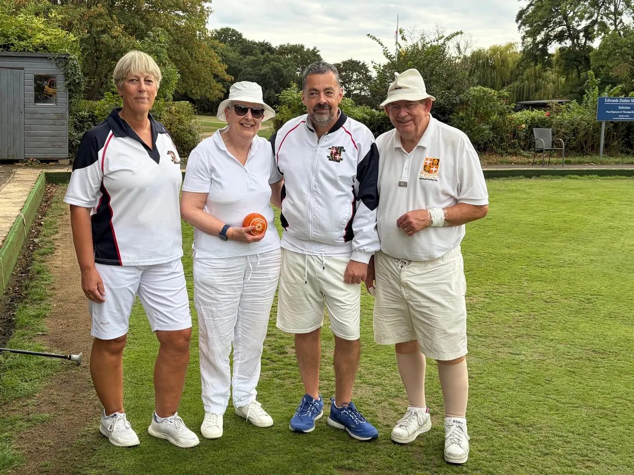 Four lawn bowls players posing together on a grass green at a bowling club, wearing white clothing and hats.