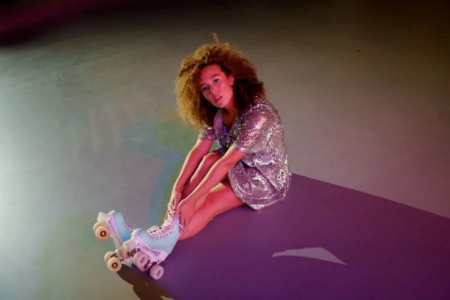 Person wearing roller skates and a sequined jacket sits on a coloured surface under atmospheric lighting.
