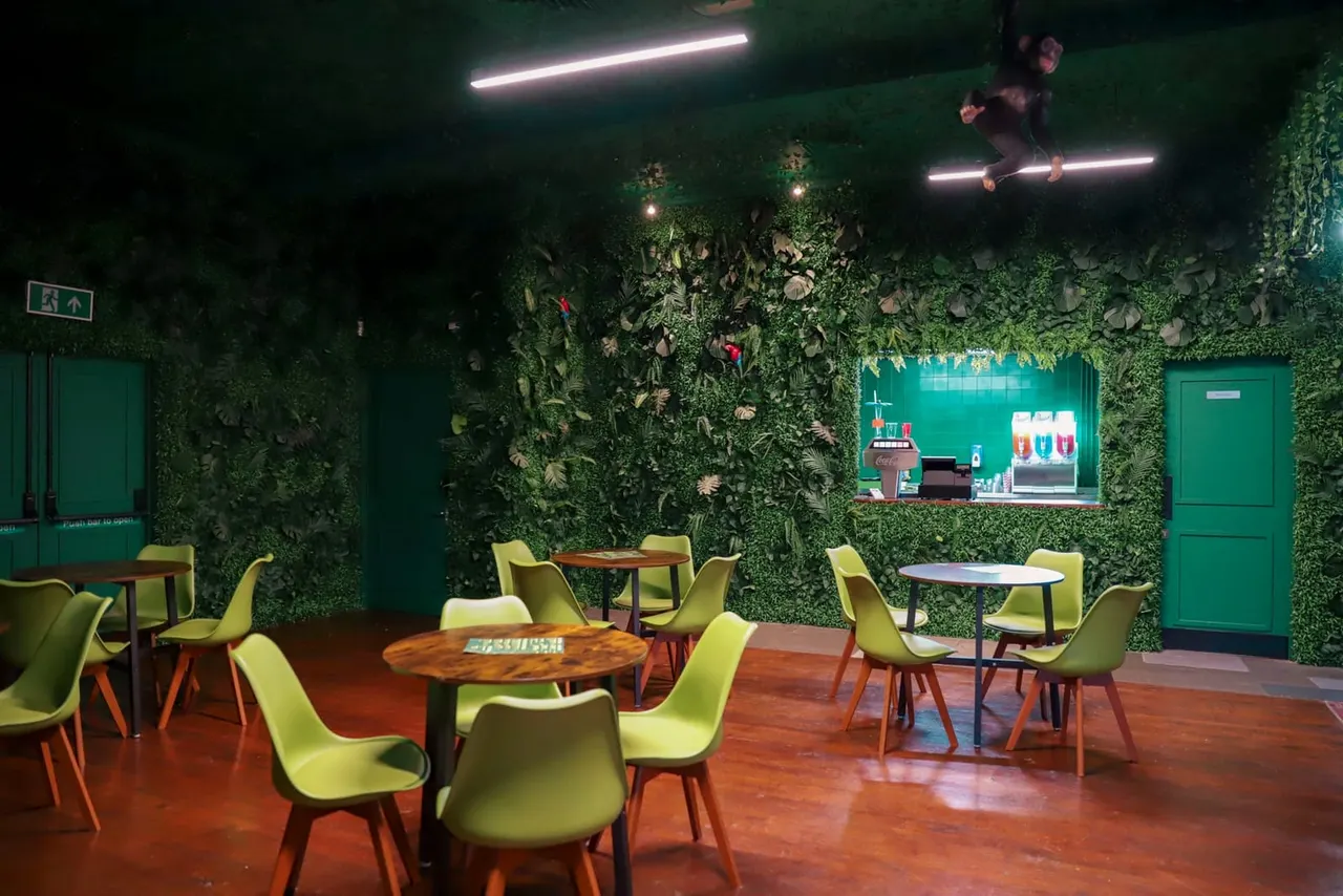 Modern indoor play space with green ivy-covered walls, lime-green chairs at wooden tables, neon lighting, and a serving counter in the background.