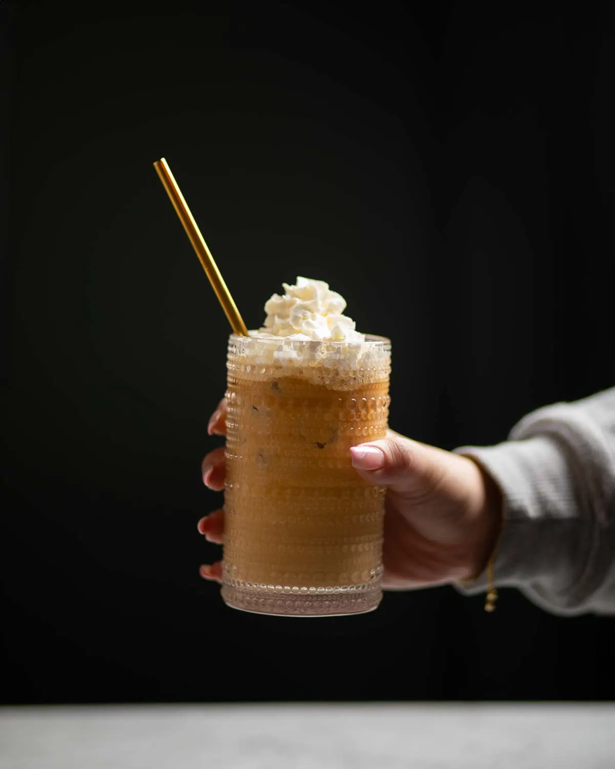 Person holding a cold coffee drink topped with whipped cream and a gold straw against a black background.