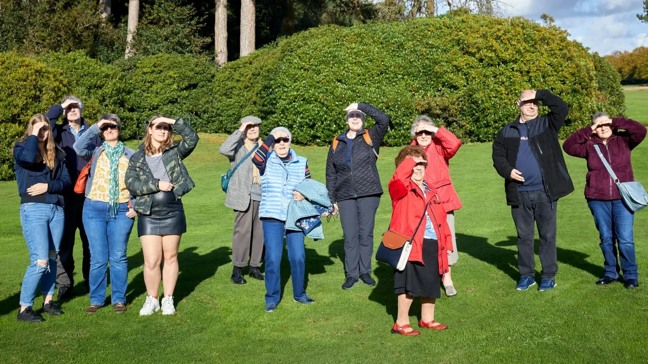 A group of visitors of various ages stand on a lawn shielding their eyes while looking upward, with manicured hedges and trees in the background.