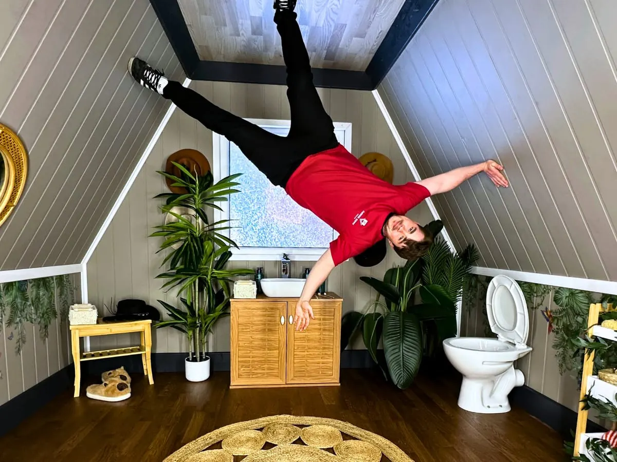Person doing a handstand in an inverted bathroom installation with toilet, sink, plants and furniture.