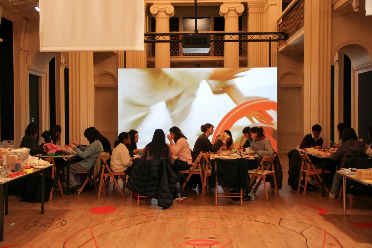 Groups of people seated at tables sharing a meal in a grand neoclassical hall with ornate columns and a large video screen.