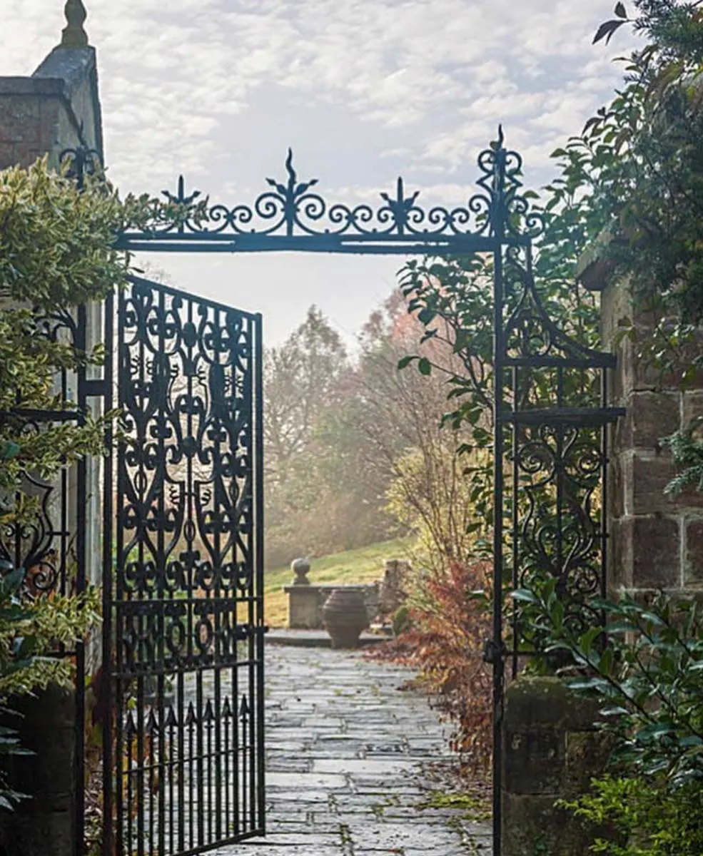 Ornate wrought-iron gate opening onto a stone pathway through a formal garden with countryside views beyond.