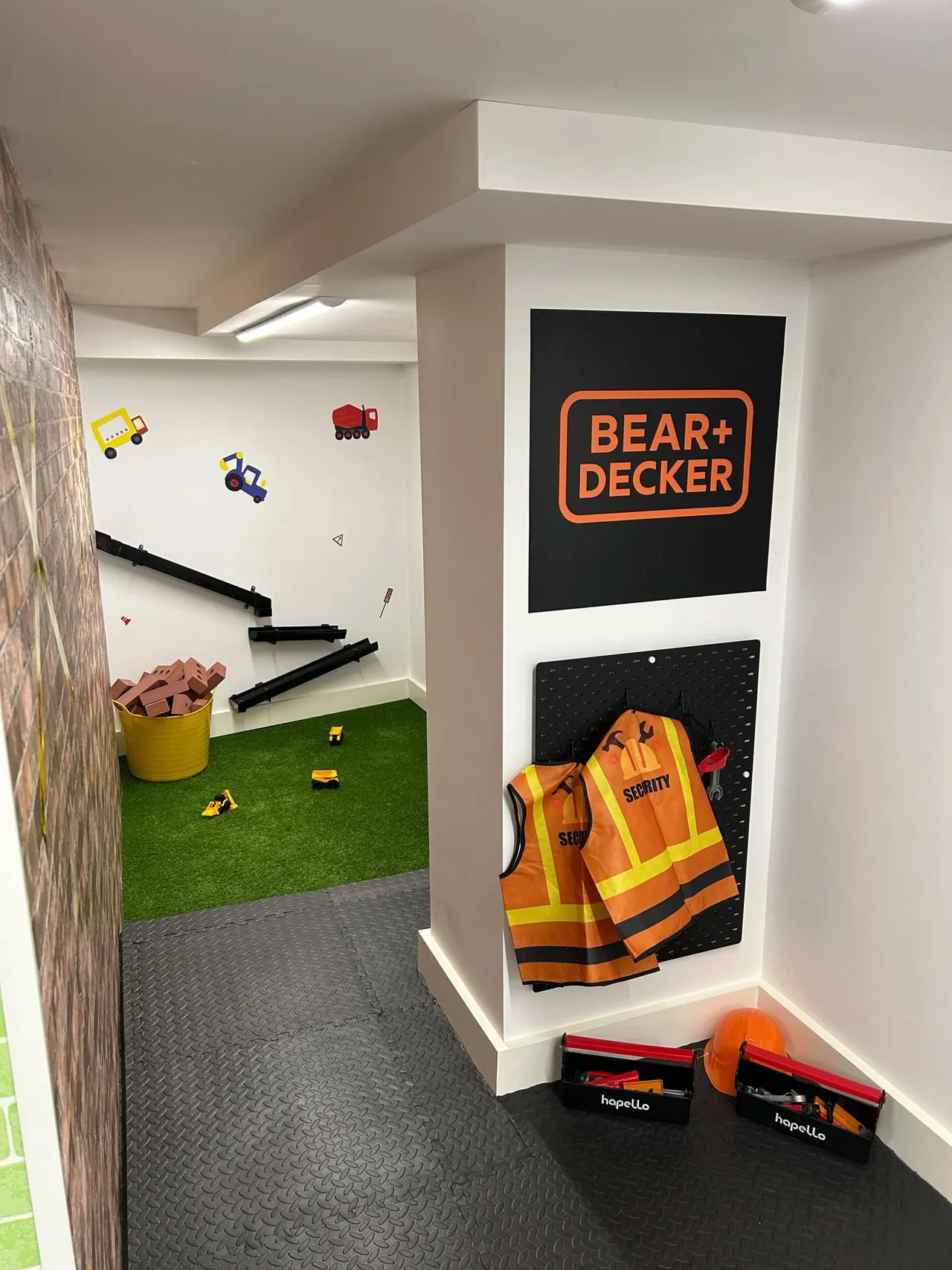 A children's play area themed around construction and building, with toy vehicles, a yellow bucket, safety vests, hard hats, and tool boxes displayed.