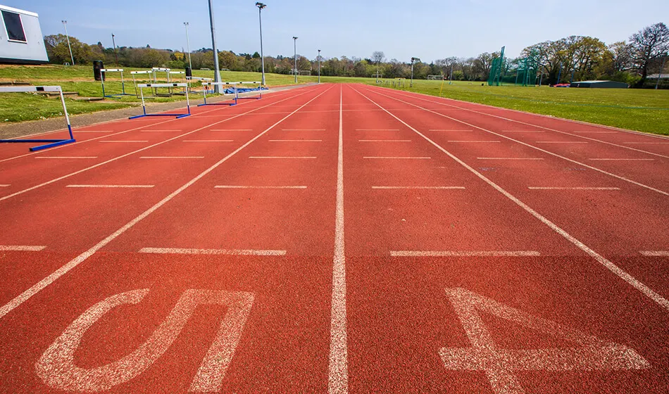 Red running track with white lane markings and numbered lanes, surrounded by green fields and sports facilities.