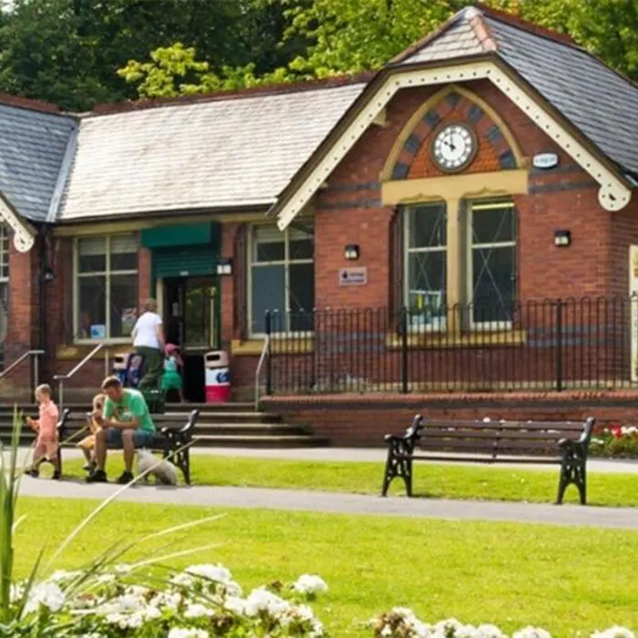 Young children and adults outside a brick community building with a clock, on a sunny lawn with benches and flowering plants.
