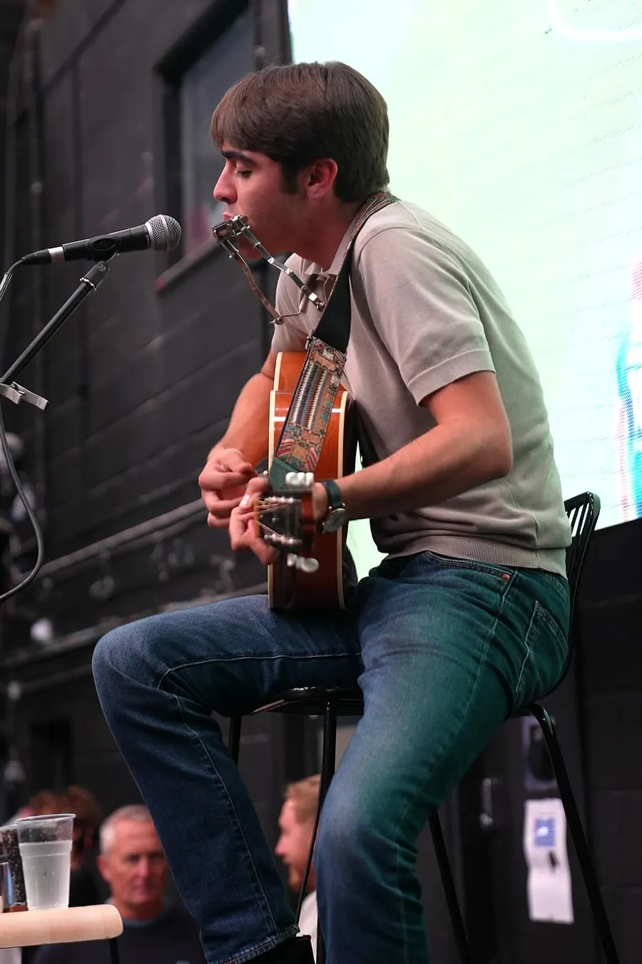 Young man sitting on a stool playing an acoustic guitar and singing into a microphone on stage.