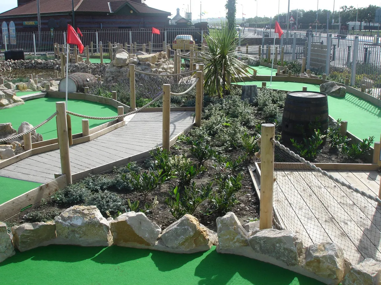 Minigolf course with bright green playing surfaces, wooden bridges, rock features, and red flags marking holes.