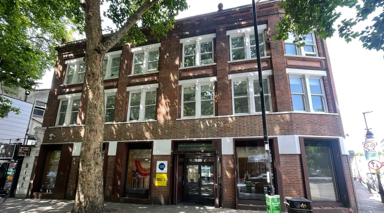 Red brick building with shop fronts at street level and residential windows above, shaded by a large tree.