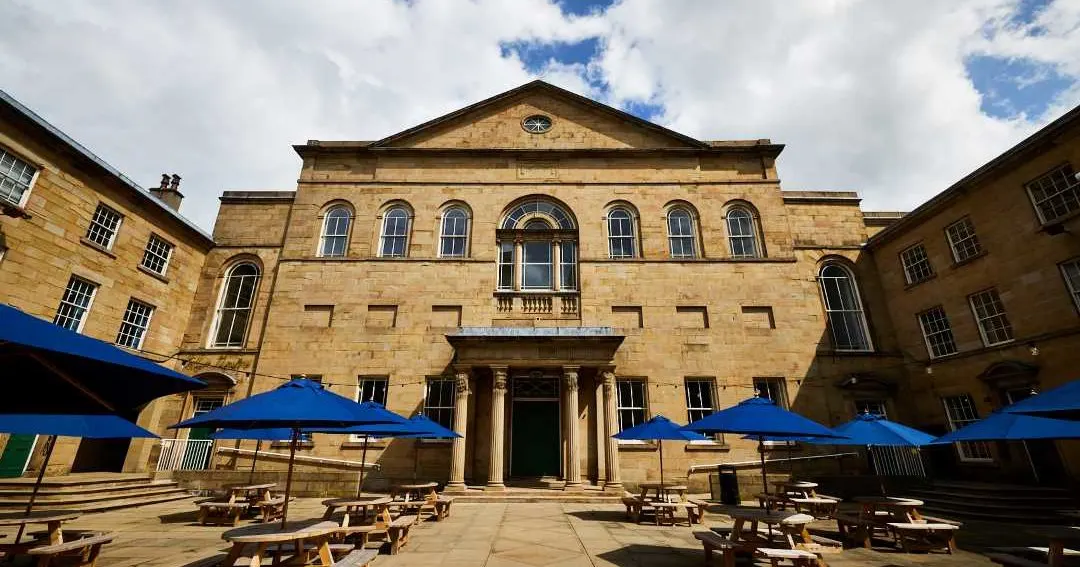 Historic stone theatre building with courtyard seating under blue umbrellas on a sunny day.