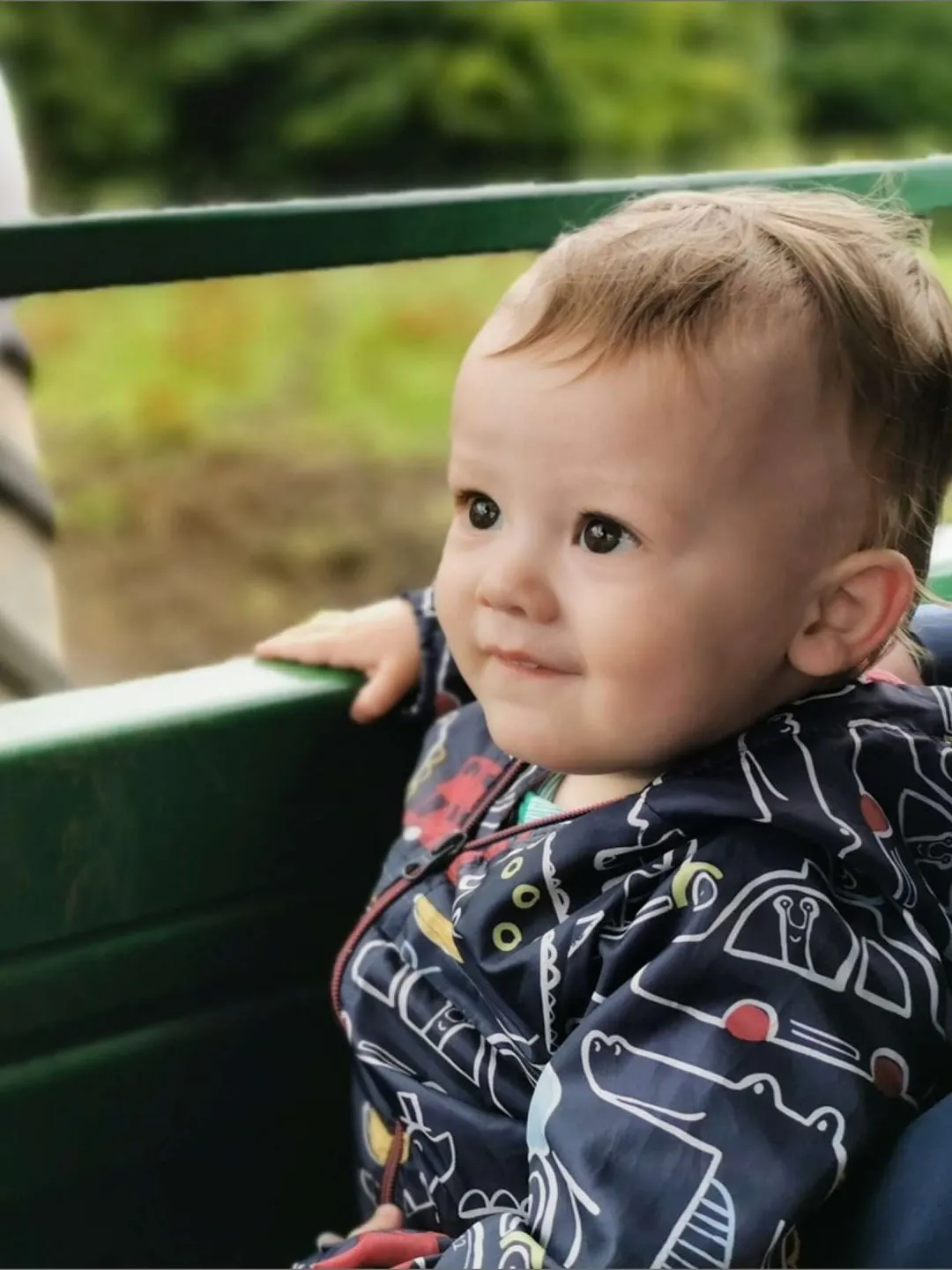 A toddler sits in a green farm vehicle, looking thoughtfully towards the camera with green fields in the background.