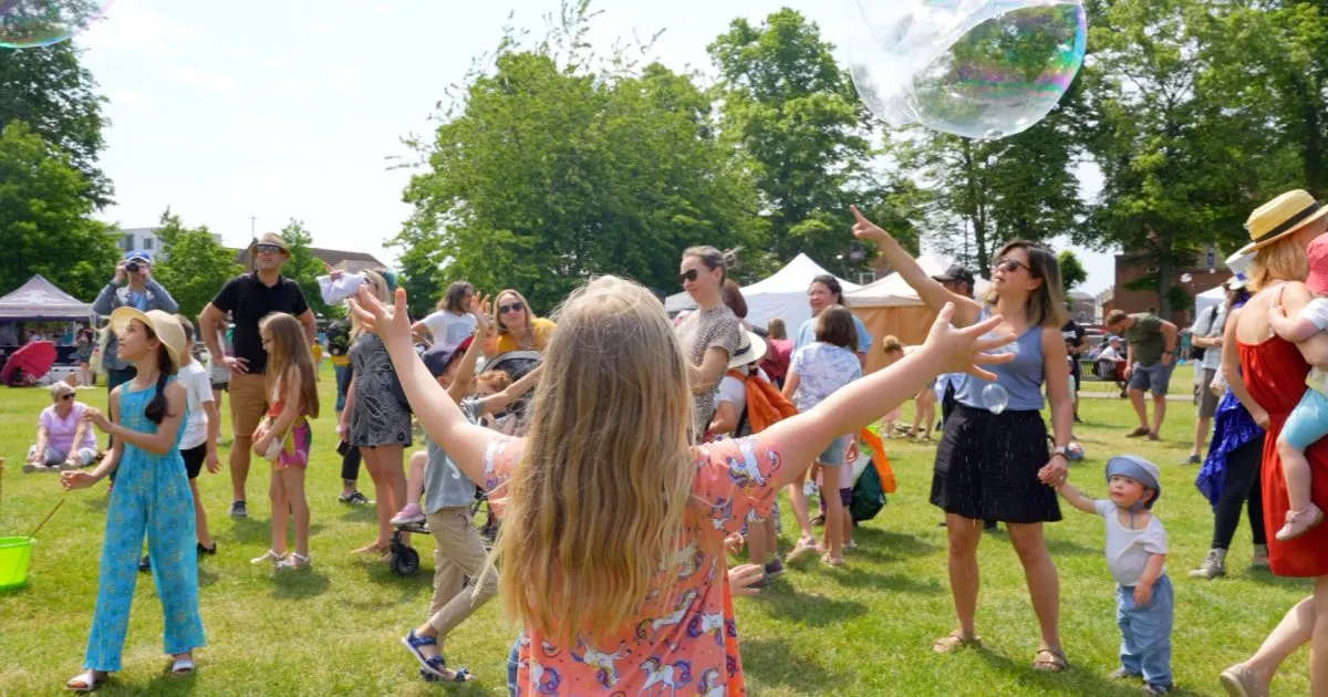 A girl with arms outstretched reaches for a giant bubble while children and adults gather on a sunny grass field at an outdoor event.