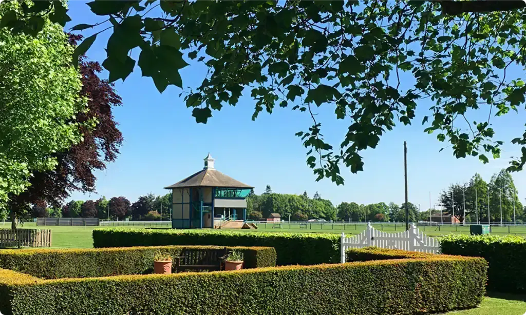 A distinctive octagonal pavilion with a thatched roof sits in a spacious manicured parkland, framed by mature trees and formal hedging.