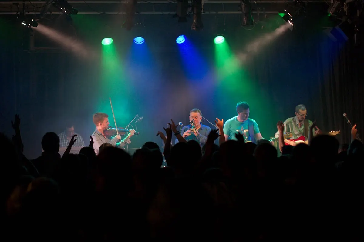 Live band performing on stage with coloured lighting, audience members with raised hands in foreground.