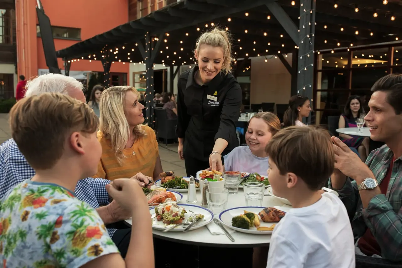 Server smiling whilst serving food to a family dining outdoors under festoon lights at an evening event.