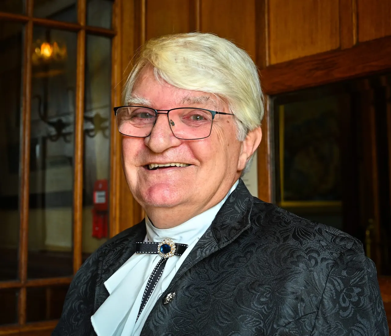 An older man with white hair and glasses wearing a dark patterned jacket and decorative bowtie, smiling indoors.