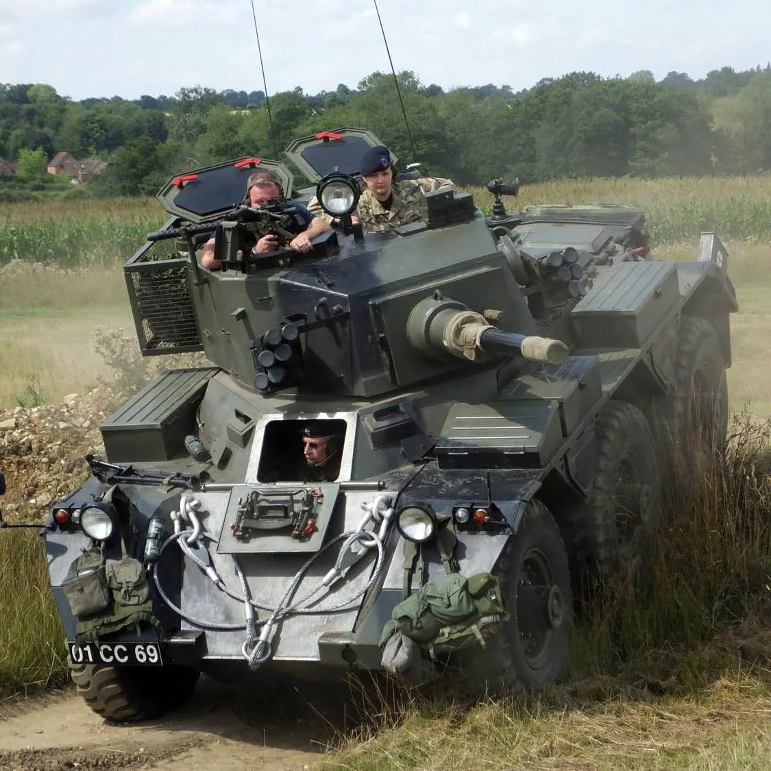 Three people operate a vintage military tank vehicle on a dusty track through countryside, with woodland and fields visible in the background.