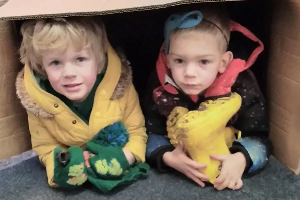 Two young children sitting inside a cardboard box, wearing colourful coats and smiling at the camera.