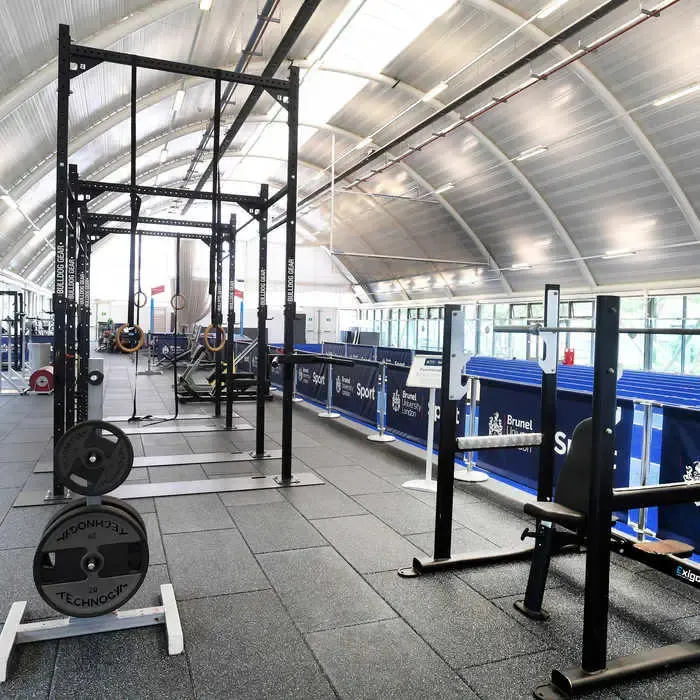 Modern gym interior with weight training equipment including squat racks and loaded barbells on a sprung floor beneath a curved metal roof.