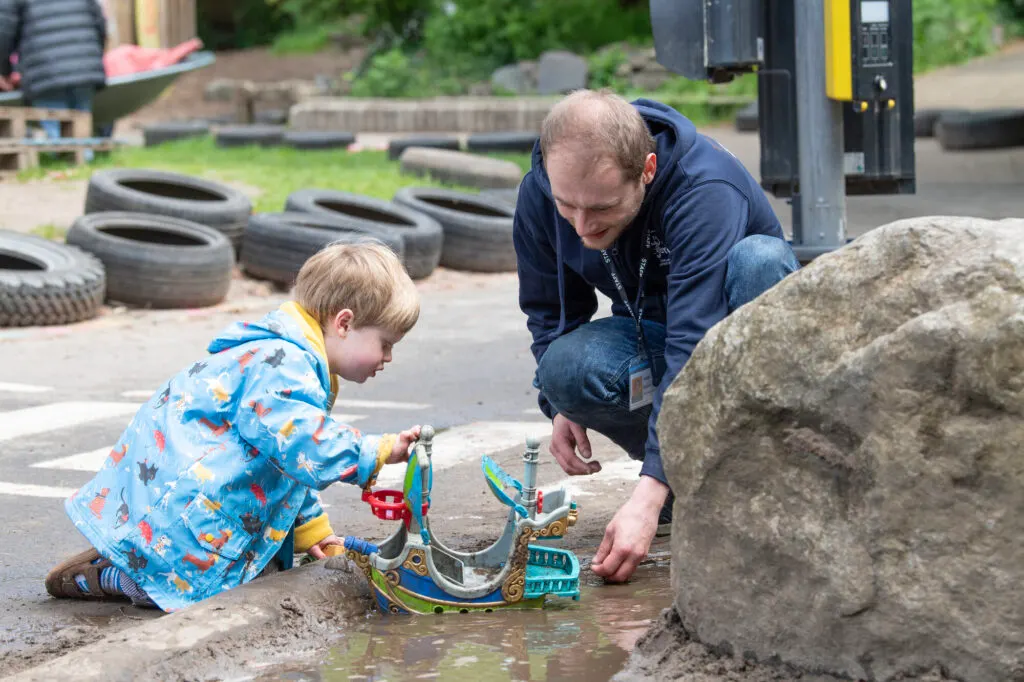 Adult staff member crouches beside young child playing with toy boats in a muddy puddle at an outdoor activity area.