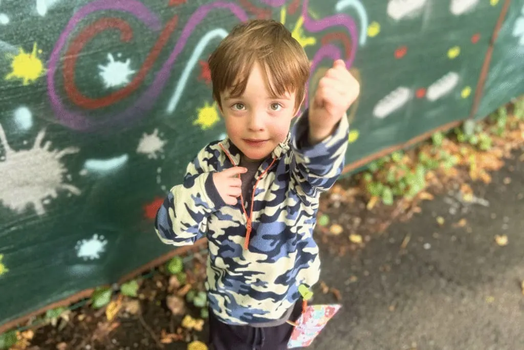 Young child in camouflage jacket pointing at camera in front of colourful chalk drawing on green board.