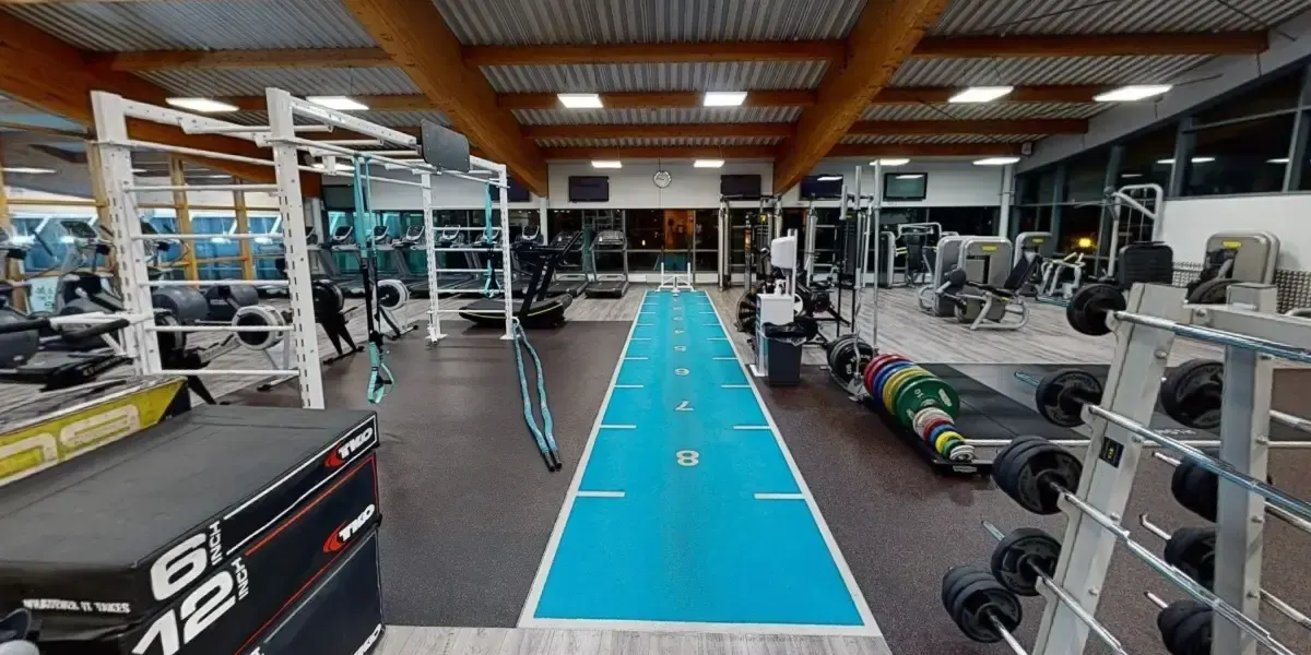 Modern gymnasium with blue lane markings down the centre, exercise equipment and weights racks lining the sides, and people using various machines.