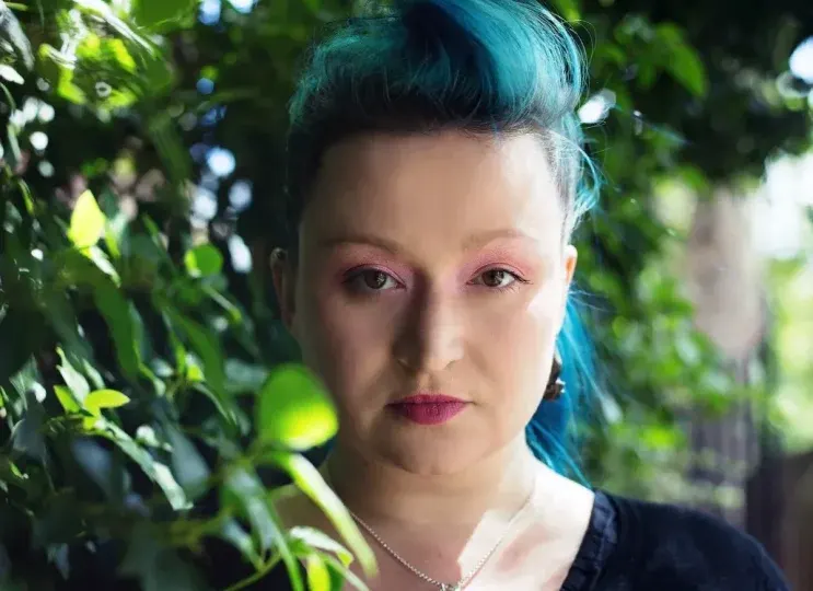 Person with blue hair and pink eyeshadow posing among green foliage outdoors.