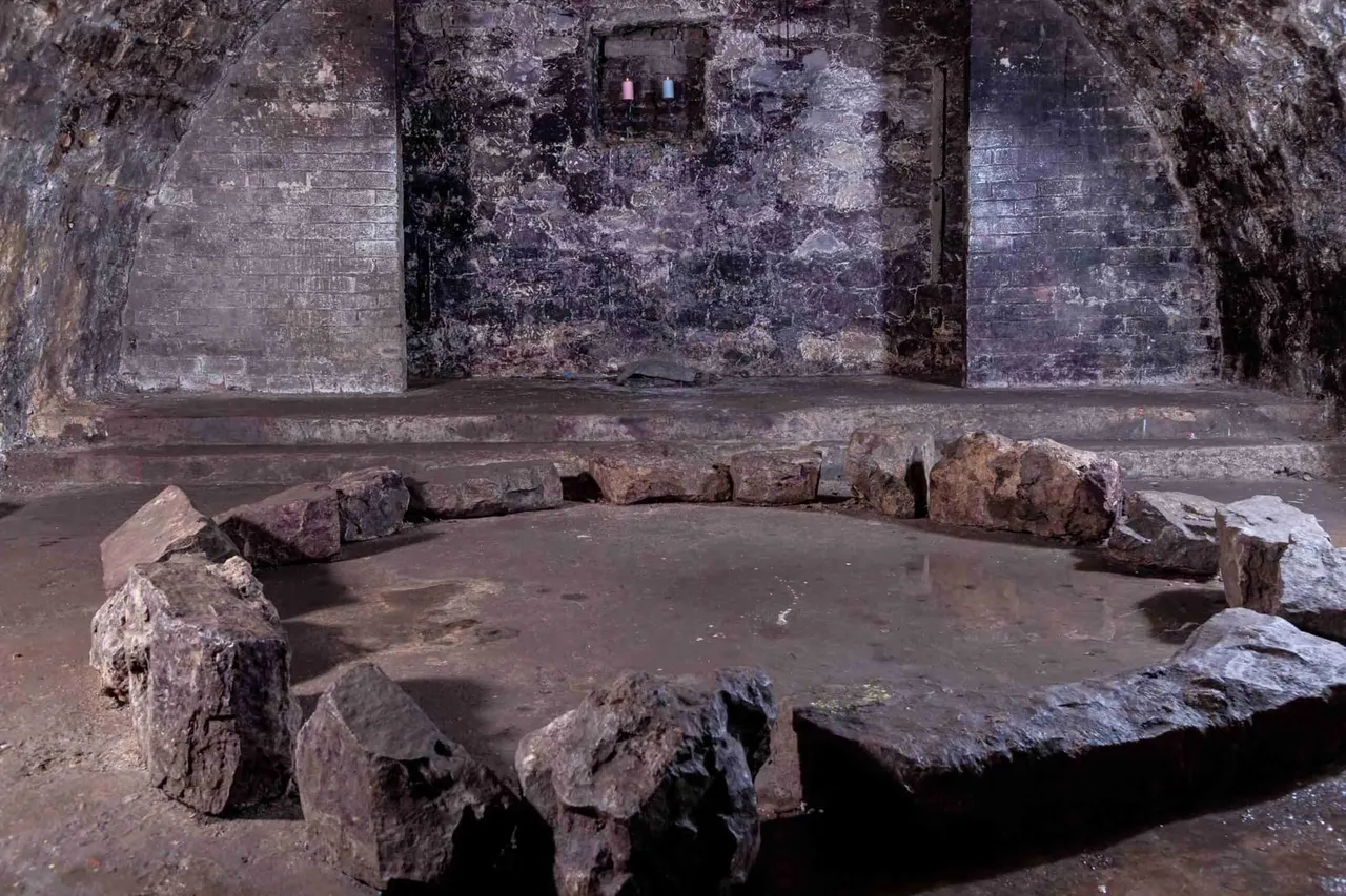 Ancient stone seating arranged in a circle inside a weathered underground chamber with crumbling walls.