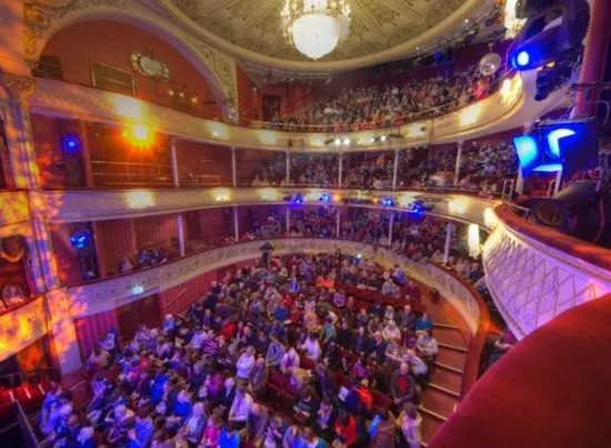 Historic theatre interior with ornate balconies and seating areas filled with audience members bathed in coloured stage lighting.