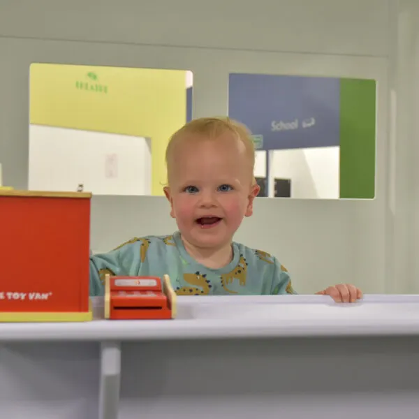A smiling toddler stands at a low table surrounded by colourful storage boxes and a toy vehicle.