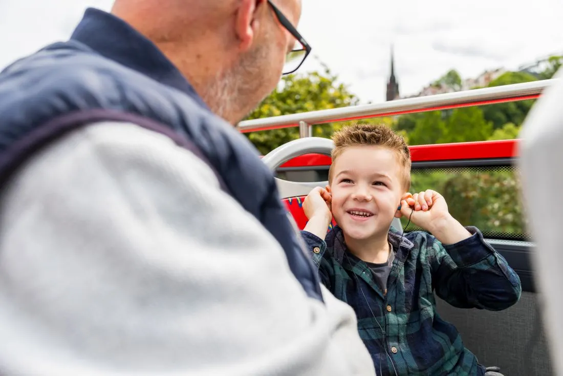 Adult and young child smiling together on an open-top tour bus with city views behind them.