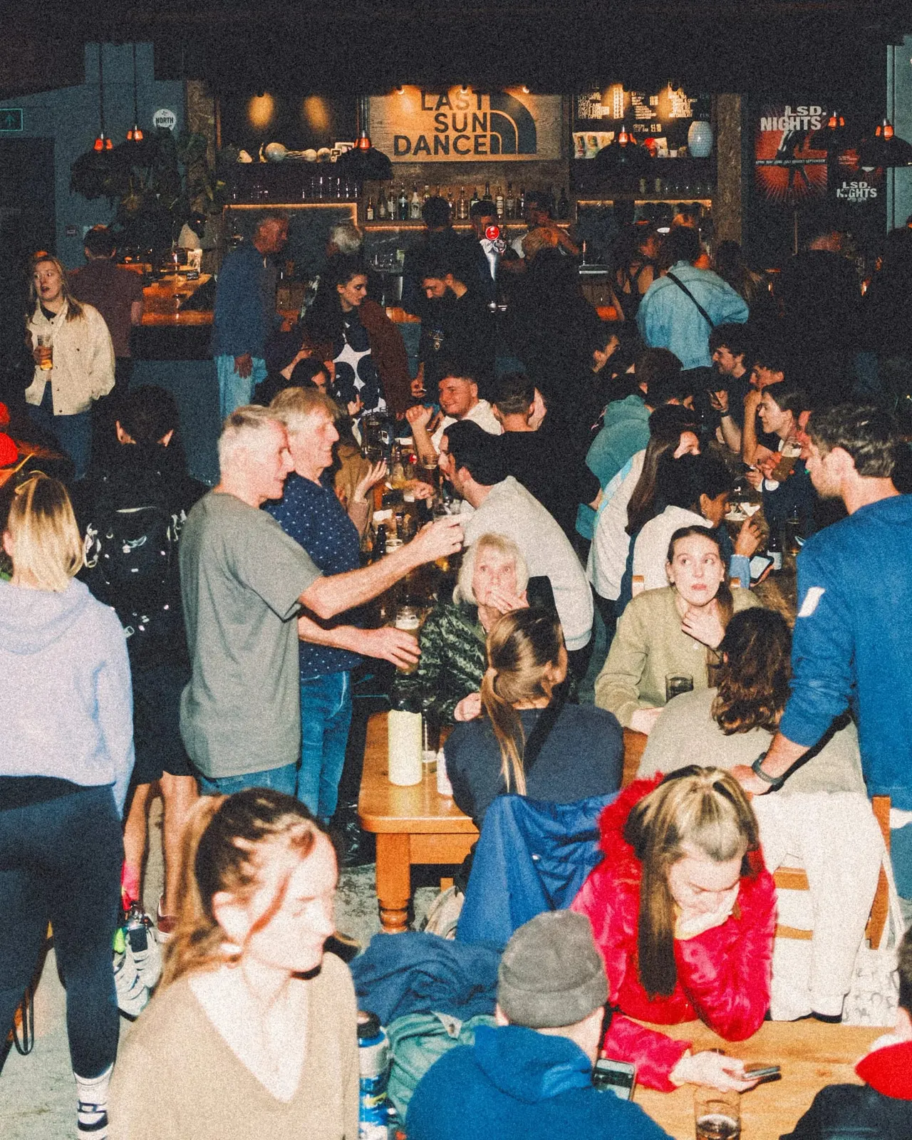 Crowded indoor bar or nightclub with patrons drinking and socialising, bar counter visible in background with illuminated signage.